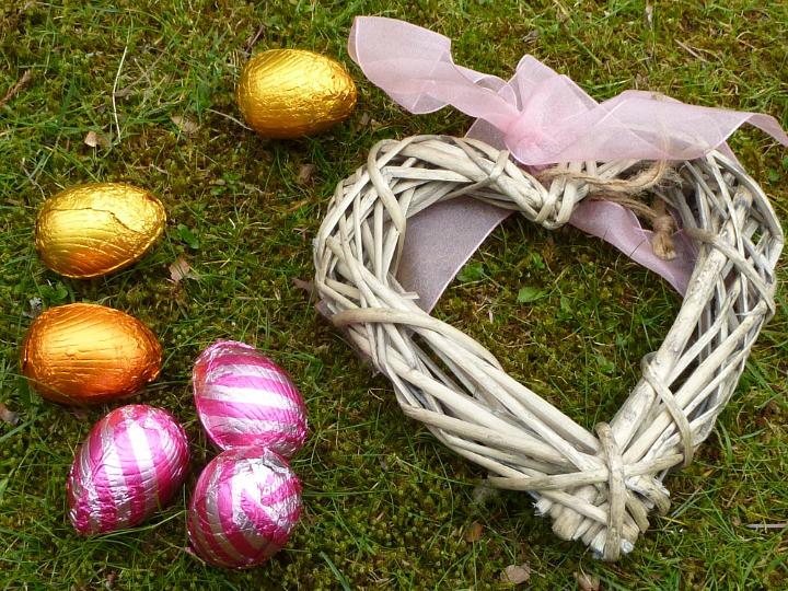 chocolate_love.jpg - Chocolate Easter eggs in colorful foil and wicker heart with pink bow in close-up on green lawn. Easter holiday decorations and love concept