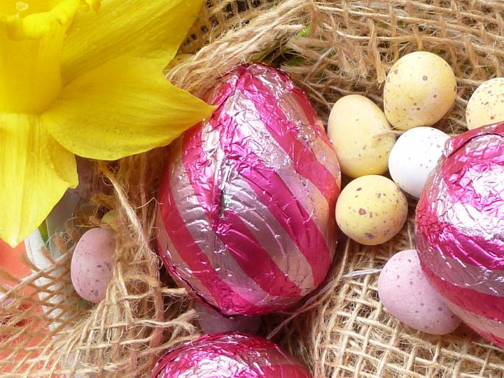easter_eggs_in_basket.jpg - Close-up of chocolate Easter eggs in striped red foil and colored chocolate quail eggs in a basket with rough sack cloth, decorated with yellow daffodil flower