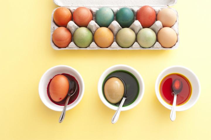 making_coloured_easter_eggs.jpg - Making homemade dyed Easter eggs with hard boiled hens eggs in pretty pastel colors with cups of dye on a yellow background viewed from above and finished eggs in a carton