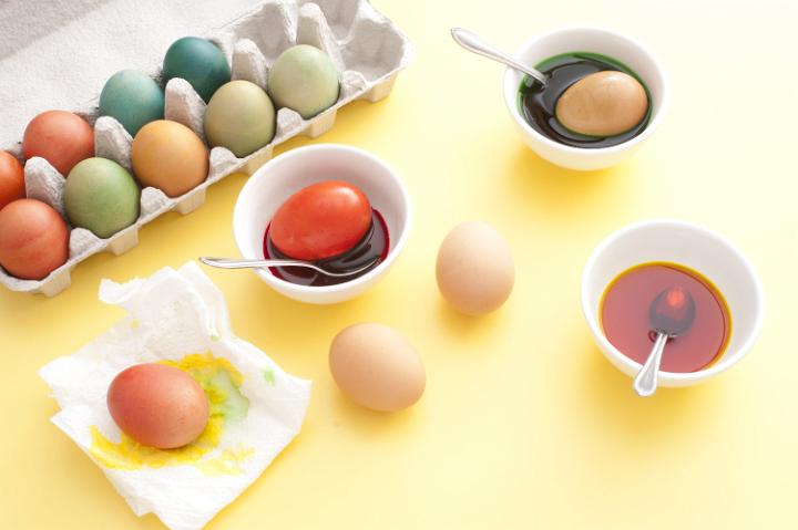 pace_egg_colouring.jpg - From above view of several painted Easter eggs in box and process of dyeing with different colors using bowls and spoons, over yellow table background
